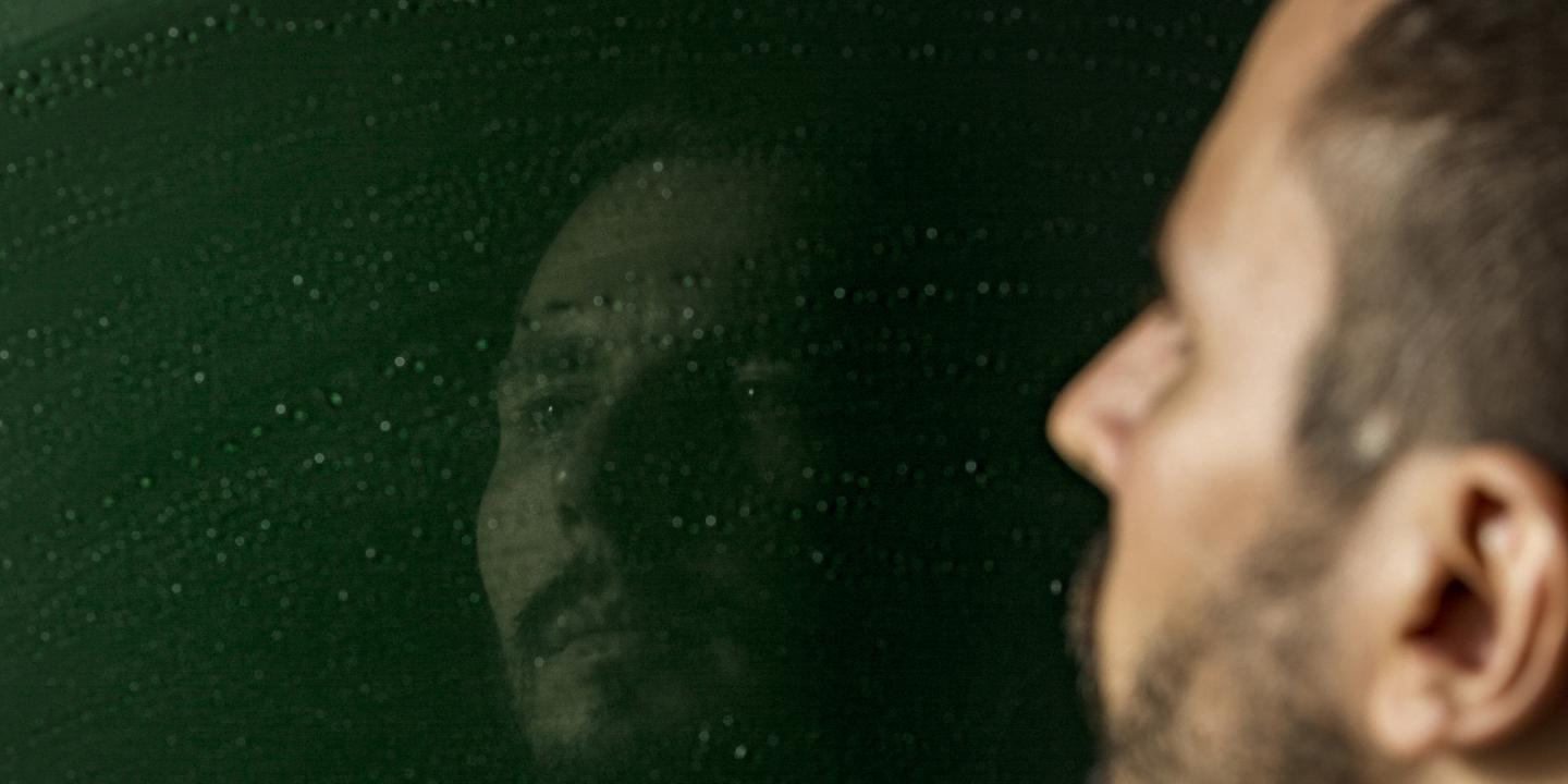 Vīrietis raugās savā atspulgā uz tumši zaļas virsmas ar gaismas atspīdumiem.