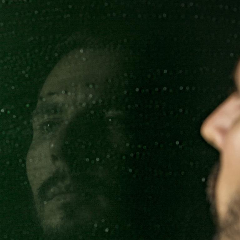 Vīrietis raugās savā atspulgā uz tumši zaļas virsmas ar gaismas atspīdumiem.