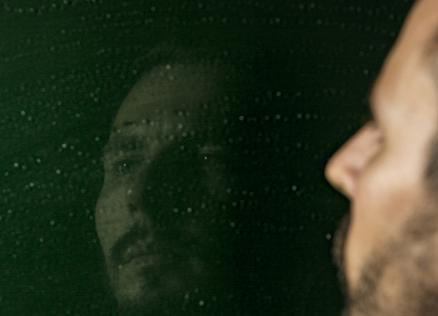 Vīrietis raugās savā atspulgā uz tumši zaļas virsmas ar gaismas atspīdumiem.