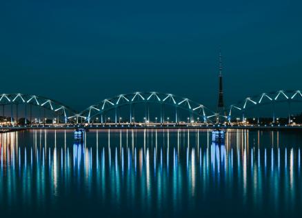 Nakts ainava ar apgaismotu tiltu pār upi un televīzijas torni fonā, atspulgi ūdenī.