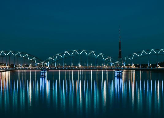 Nakts ainava ar apgaismotu tiltu pār upi un televīzijas torni fonā, atspulgi ūdenī.