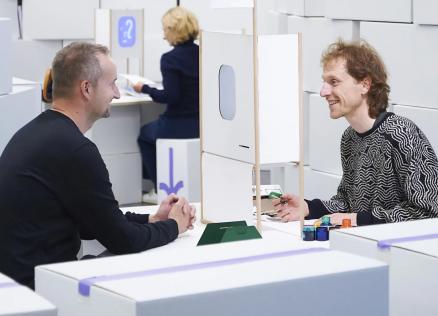 Divi vīrieši sēž pie galda un sarunājas, starp viņiem novietota neliela konstrukcija, fonā redzama vēl viena persona.