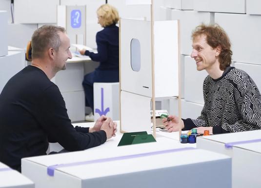 Divi vīrieši sēž pie galda un sarunājas, starp viņiem novietota neliela konstrukcija, fonā redzama vēl viena persona.