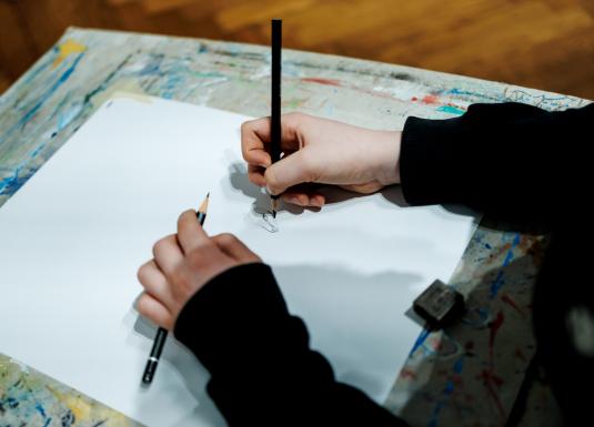 Rokas ar zīmuļiem skicē zīmējumu uz baltas lapas uz krāsām notraipīta galda.