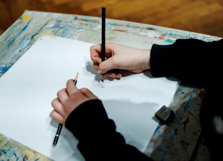 Rokas ar zīmuļiem skicē zīmējumu uz baltas lapas uz krāsām notraipīta galda.