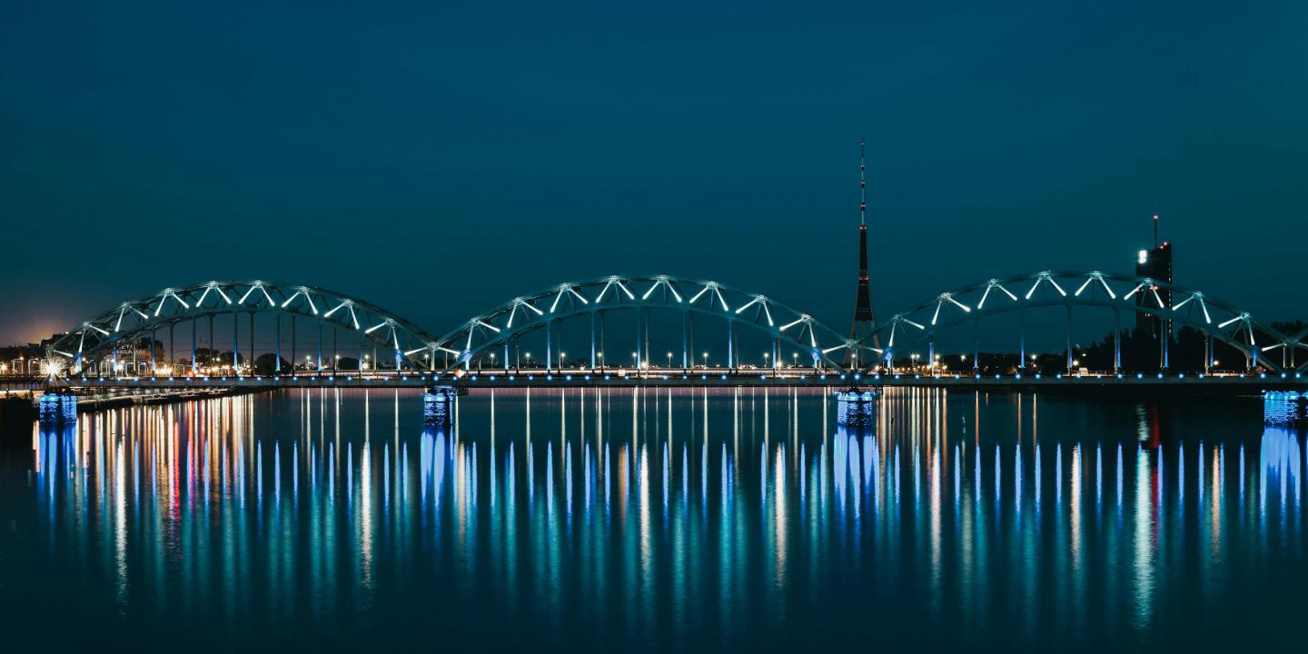 Nakts ainava ar apgaismotu tiltu pār upi un televīzijas torni fonā, atspulgi ūdenī.