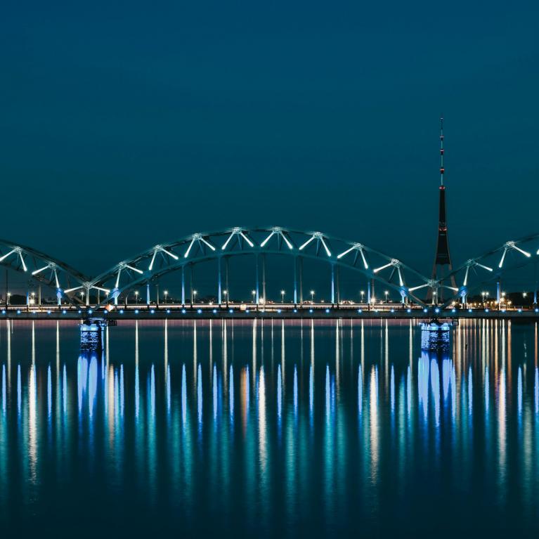 Nakts ainava ar apgaismotu tiltu pār upi un televīzijas torni fonā, atspulgi ūdenī.