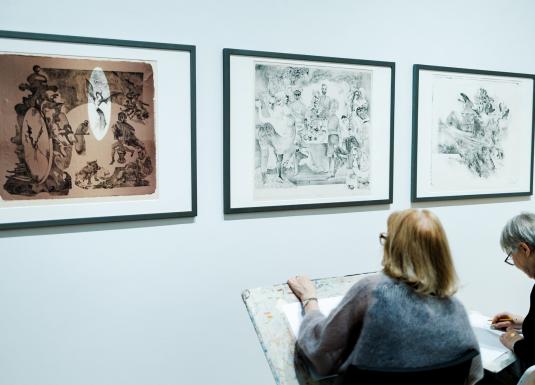 Divas sievietes muzejā sēž un skicē, vērojot pie sienas izstādītus grafikas darbus.