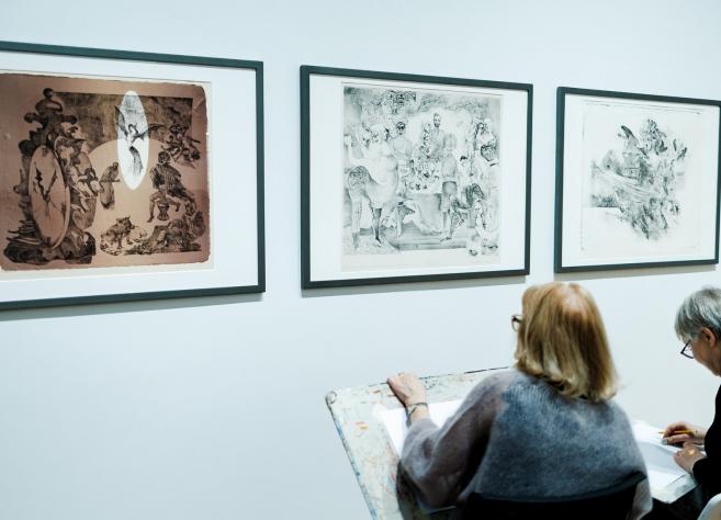 Divas sievietes muzejā sēž un skicē, vērojot pie sienas izstādītus grafikas darbus.