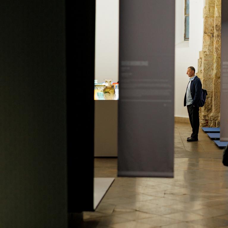 Muzeja zālē apmeklētāji apskata ekspozīciju; priekšplānā daļēji redzams dekoratīvs krēsls, fonā vitrīna ar stikla traukiem un uz pjedestāla novietots stikla galdiņš.