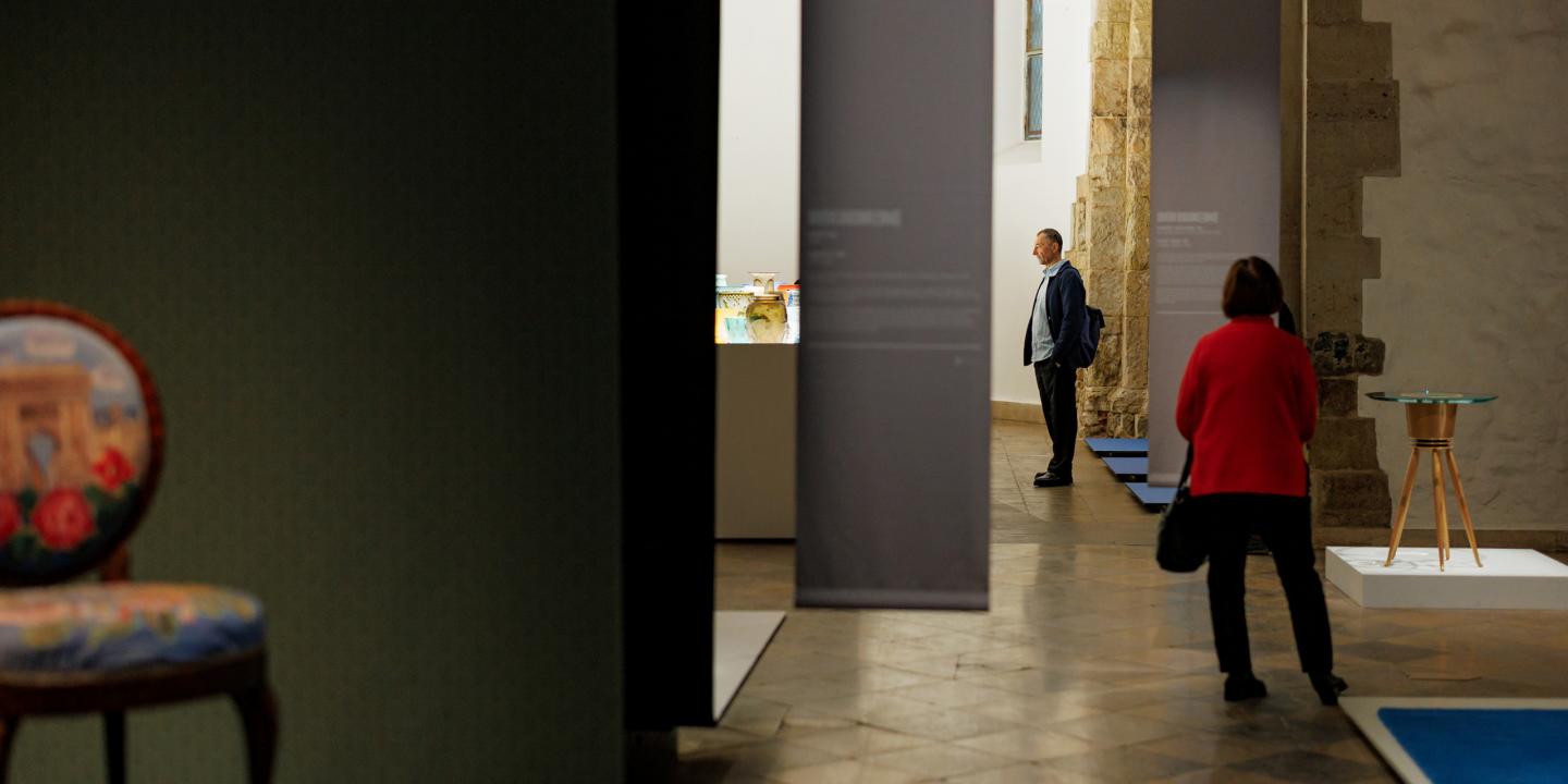 Muzeja zālē apmeklētāji apskata ekspozīciju; priekšplānā daļēji redzams dekoratīvs krēsls, fonā vitrīna ar stikla traukiem un uz pjedestāla novietots stikla galdiņš.