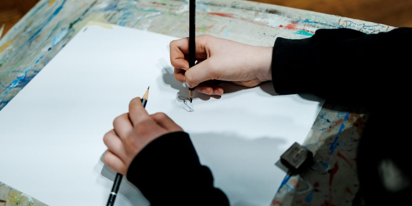 Rokas ar zīmuļiem skicē zīmējumu uz baltas lapas uz krāsām notraipīta galda.