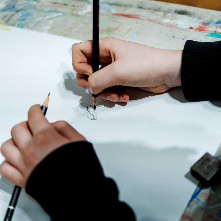 Rokas ar zīmuļiem skicē zīmējumu uz baltas lapas uz krāsām notraipīta galda.