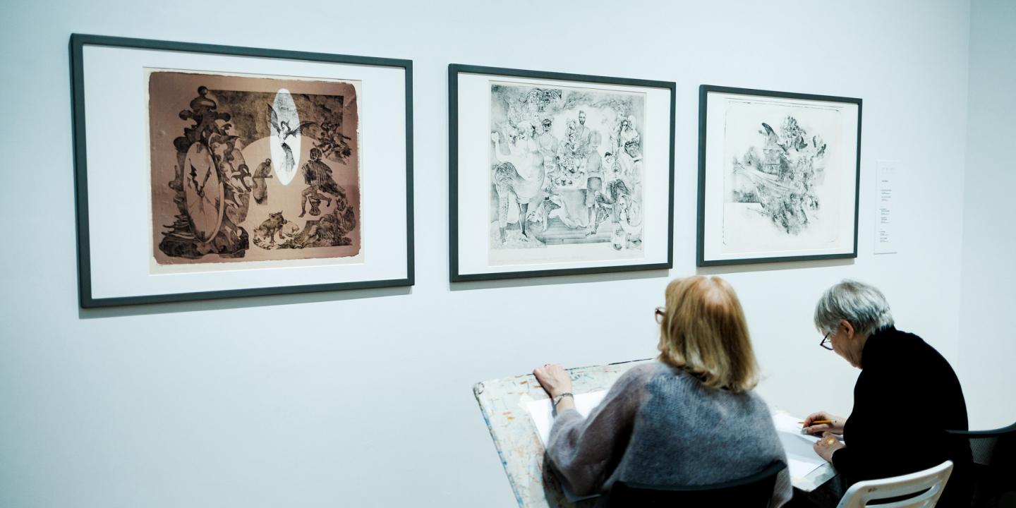 Divas sievietes muzejā sēž un skicē, vērojot pie sienas izstādītus grafikas darbus.