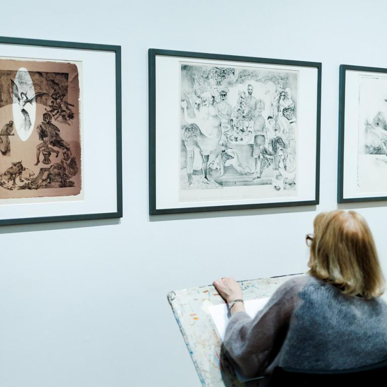 Divas sievietes muzejā sēž un skicē, vērojot pie sienas izstādītus grafikas darbus.