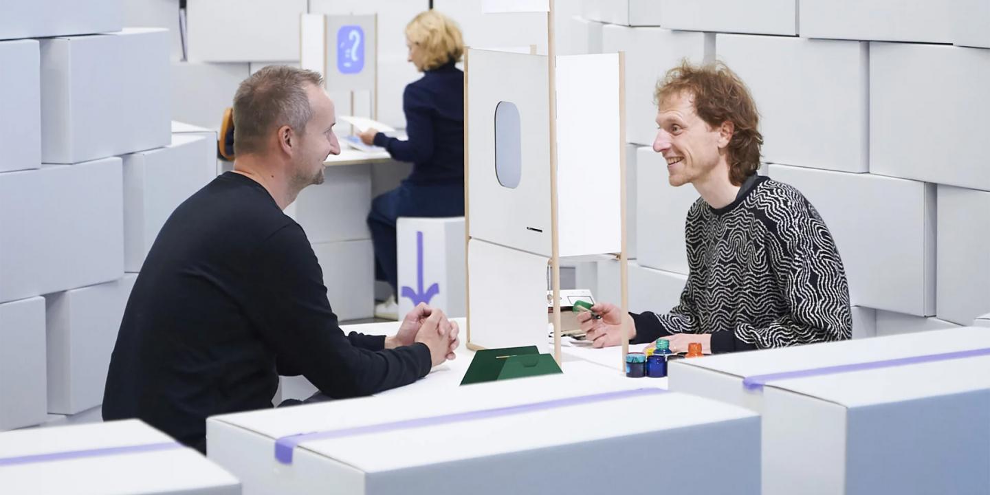 Divi vīrieši sēž pie galda un sarunājas, starp viņiem novietota neliela konstrukcija, fonā redzama vēl viena persona.