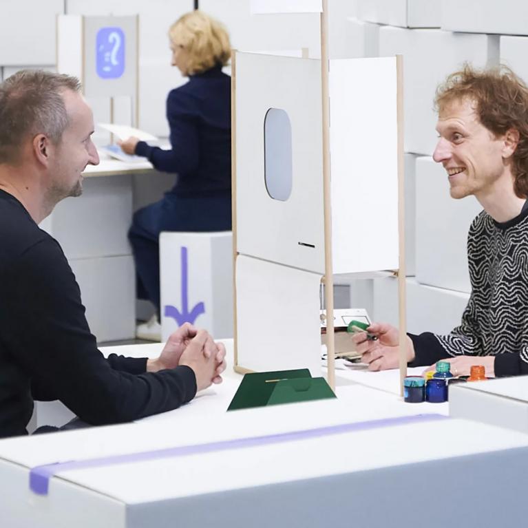 Divi vīrieši sēž pie galda un sarunājas, starp viņiem novietota neliela konstrukcija, fonā redzama vēl viena persona.