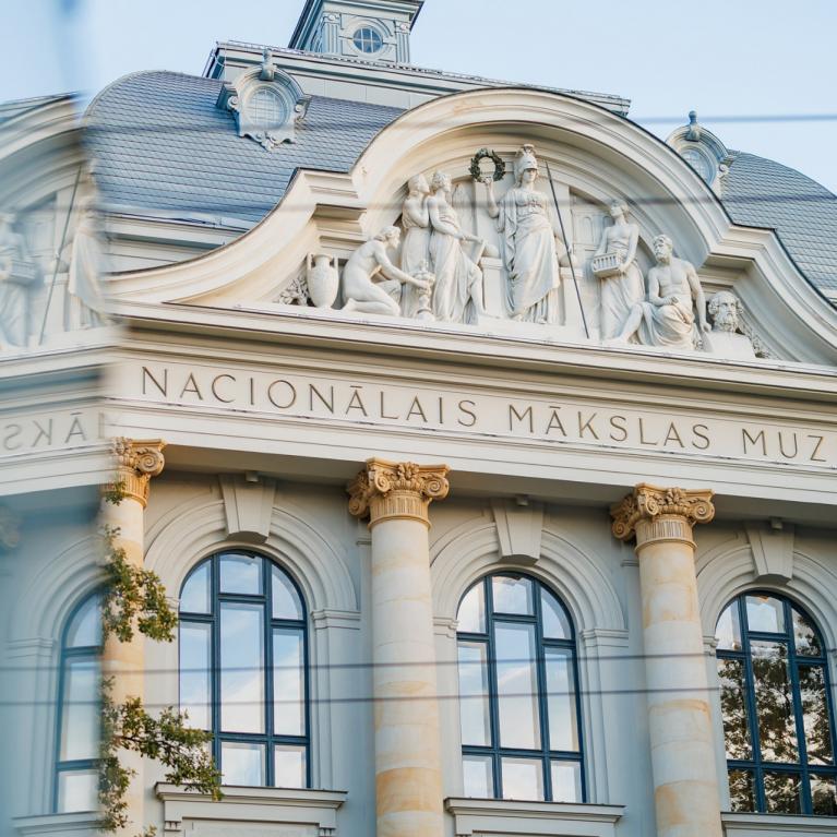 Latvijas Nacionālā mākslas muzeja fasāde ar klasiskām kolonnām un skulptūrām, redzama aiz stikla atspīduma.