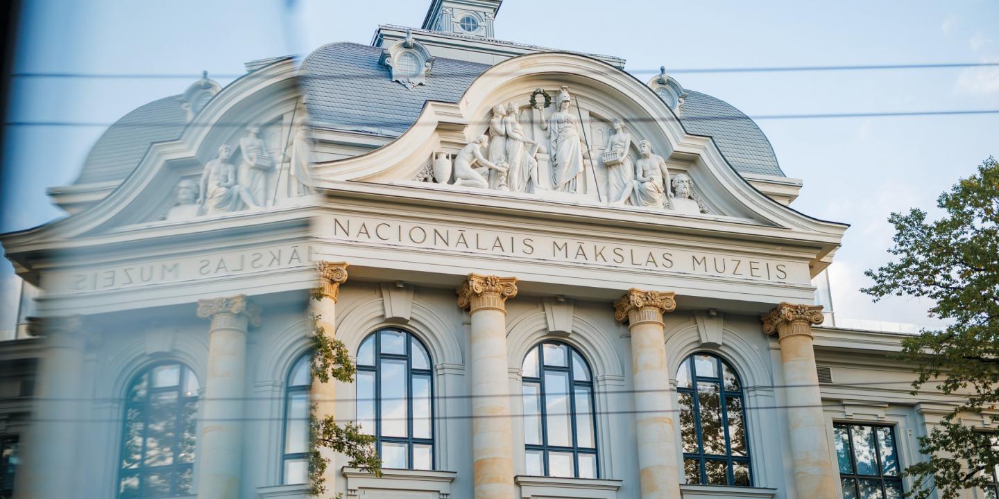 Latvijas Nacionālā mākslas muzeja fasāde ar klasiskām kolonnām un skulptūrām, redzama aiz stikla atspīduma.