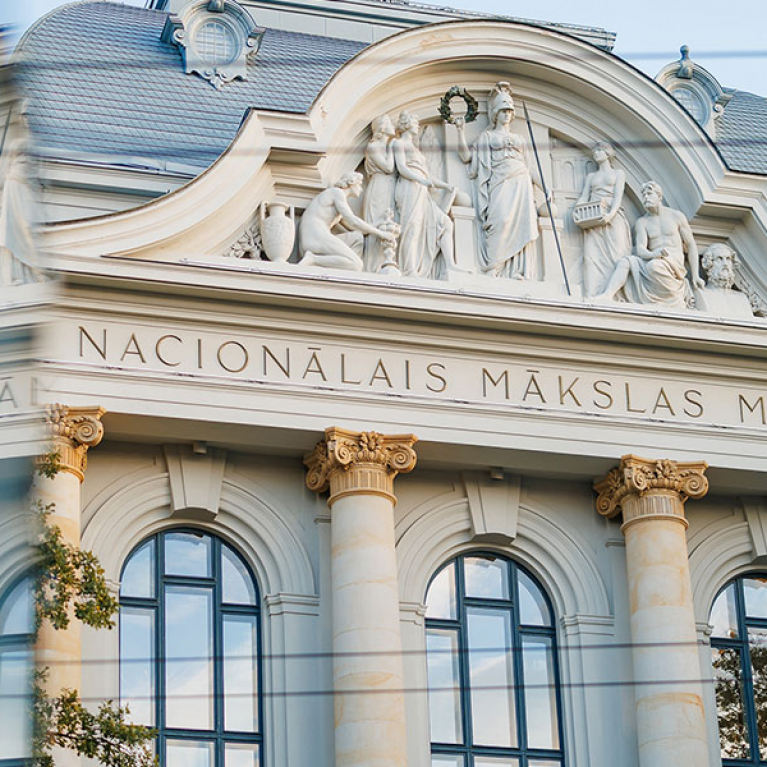 Latvijas Nacionālā mākslas muzeja fasāde ar klasiskām kolonnām un skulptūrām, redzama aiz stikla atspīduma.