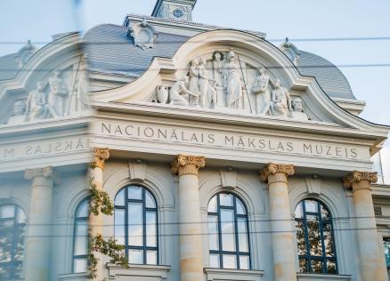 Latvijas Nacionālā mākslas muzeja fasāde ar klasiskām kolonnām un skulptūrām, redzama aiz stikla atspīduma.