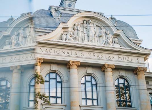 Latvijas Nacionālā mākslas muzeja fasāde ar klasiskām kolonnām un skulptūrām, redzama aiz stikla atspīduma.