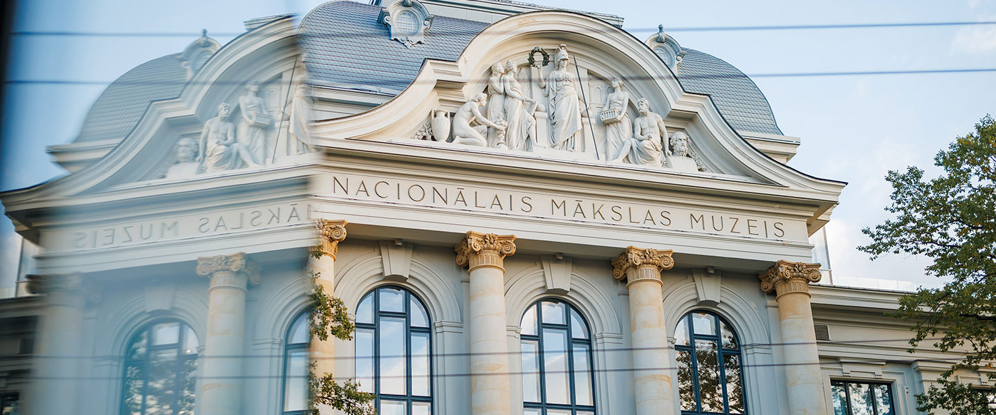 The façade of the Latvian National Museum of Art with classical columns and sculptures, seen through a glass reflection.