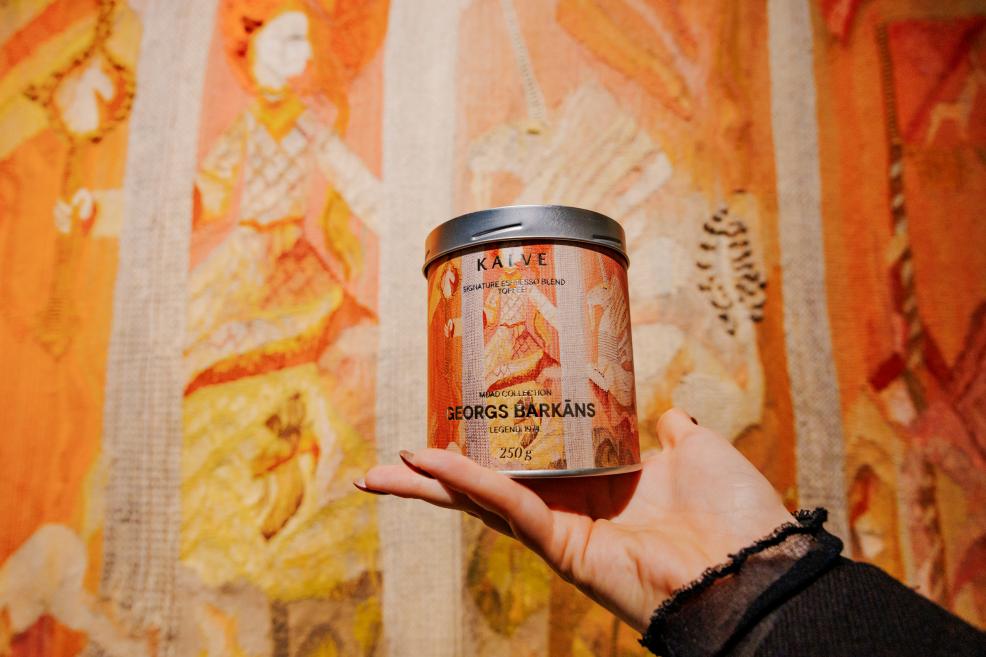 Rokā turēta kafijas bundža KALVE – Georgs Barkāns ar oranži tonētu etiķeti, fotografēta uz dekoratīvas gobelēna tekstilijas fona siltos toņos.