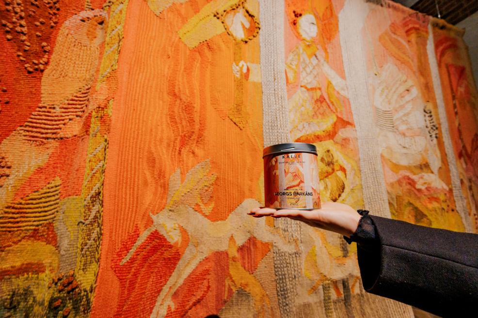 Rokā turēta kafijas bundža KALVE – Georgs Barkāns ar oranži tonētu etiķeti, fotografēta uz dekoratīvas gobelēna tekstilijas fona siltos toņos.