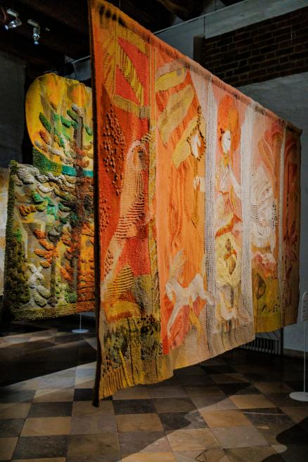 Telpā pie griestiem piekārts liela formāta gobelēns siltos oranžos un okera toņos ar figurāliem un simboliskiem motīviem, skatā no sāniem muzeja ekspozīcijā.