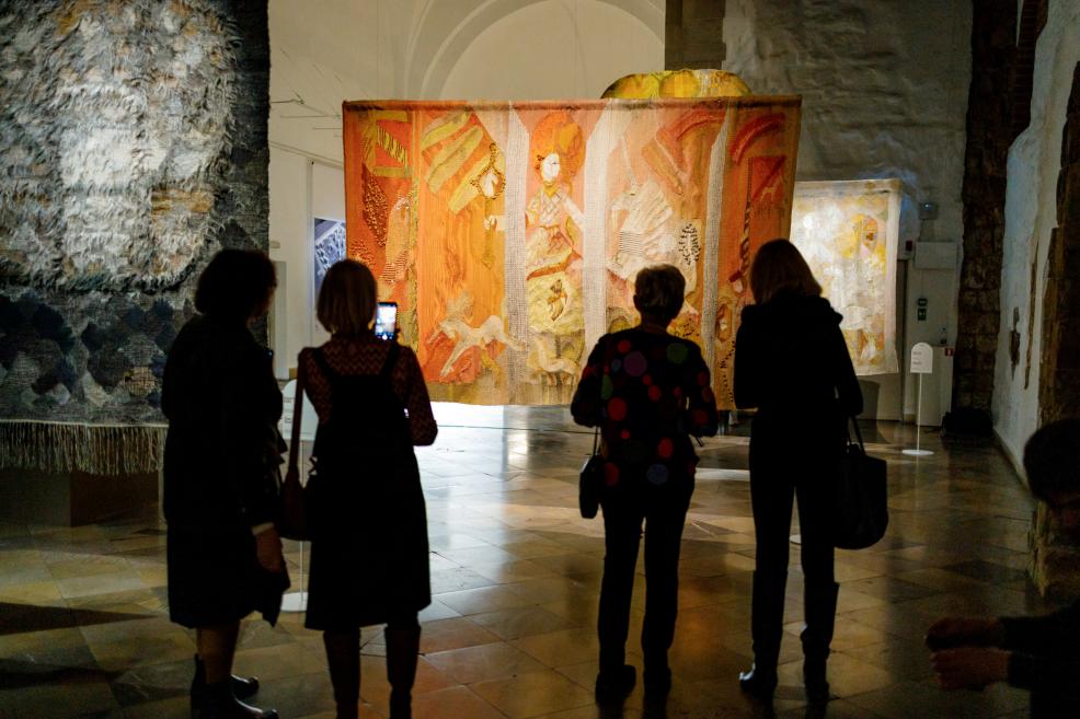 Muzeja apmeklētāji stāv ekspozīcijas telpā un vēro pie griestiem piekārtu liela formāta gobelēnu siltos oranžos toņos, kas izgaismots plašā akmens velvju zālē.