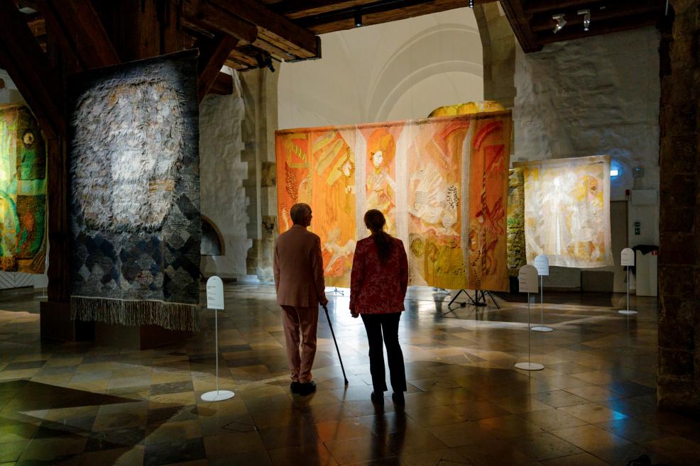 Muzeja apmeklētāji stāv ekspozīcijas telpā un vēro pie griestiem piekārtu liela formāta gobelēnu siltos oranžos toņos, kas izgaismots plašā akmens velvju zālē.