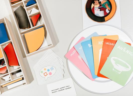 Uz galda izkārtoti krāsaini ģeometriski koka elementi kastītēs un ilustrētas kartītes ar uzrakstiem, veidojot radošu izglītojošu spēles komplektu.