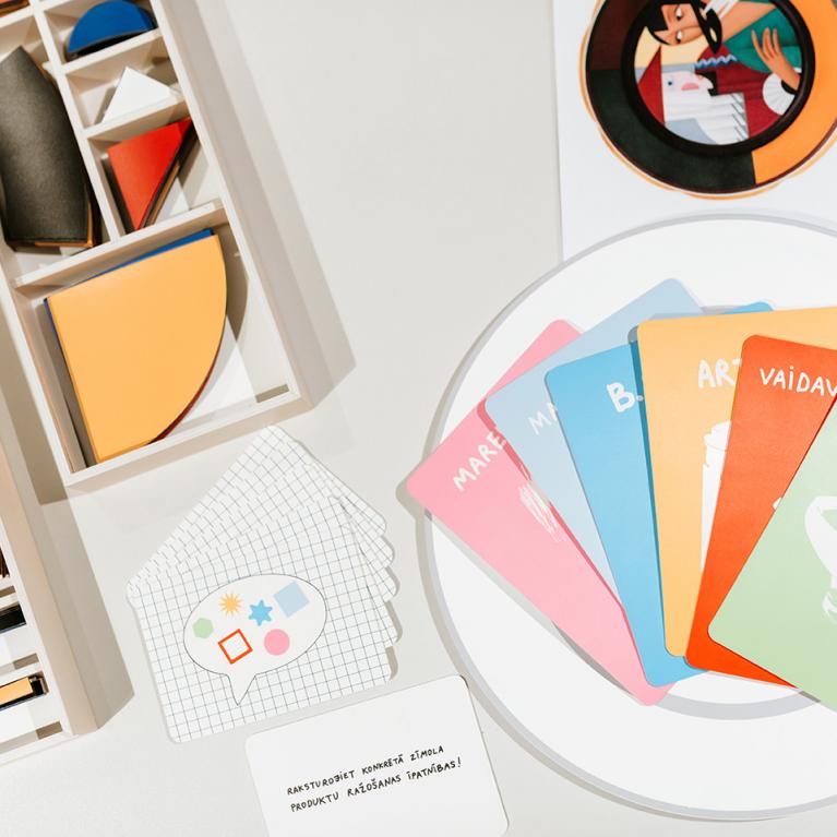 Uz galda izkārtoti krāsaini ģeometriski koka elementi kastītēs un ilustrētas kartītes ar uzrakstiem, veidojot radošu izglītojošu spēles komplektu.