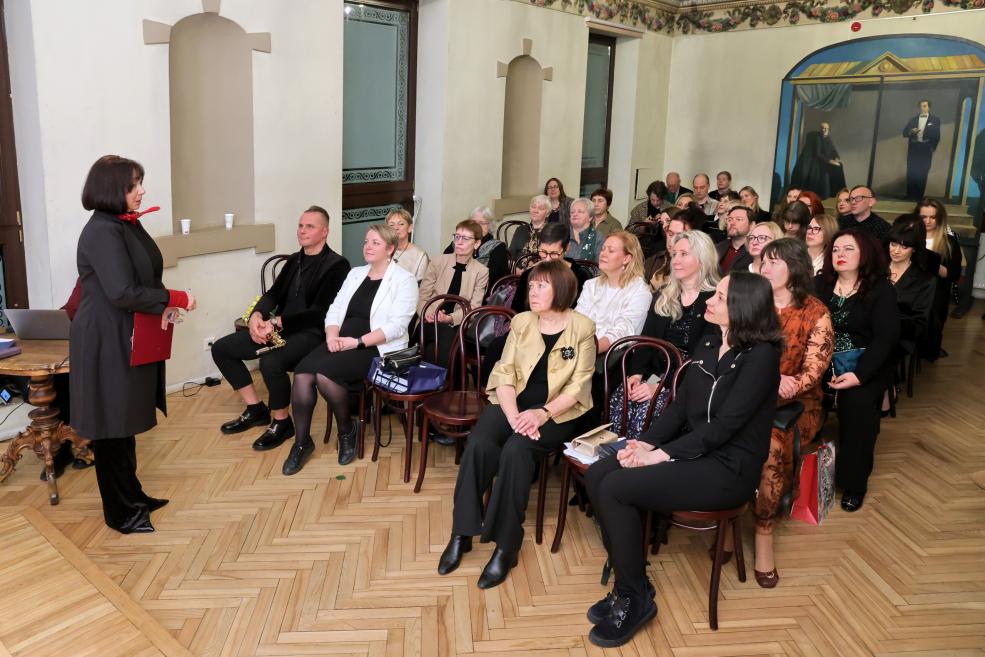 Sieviete stāv zāles priekšā un uzrunā sēdošu auditoriju ar klausītājiem vairākās rindās vēsturiskā interjerā.