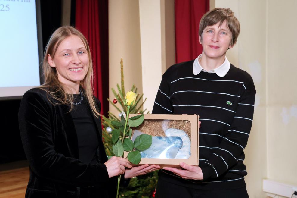 Divas sievietes stāv zālē, smaida un tur rokās dzeltenu rozi un dāvanu kastīti ar stikla objektu svinīgā pasākuma laikā.