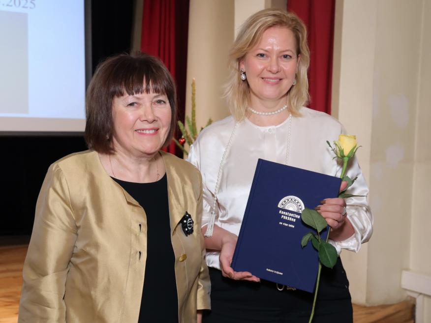 Divas sievietes stāv uz skatuves, smaida un viena no viņām tur zilu grāmatu un dzeltenu rozi svinīgā pasākumā.