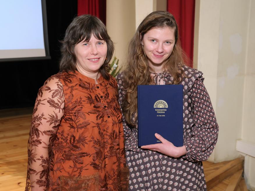 Divas sievietes stāv uz skatuves, smaida, un viena no viņām rokās tur zilu grāmatu svinīgā pasākumā