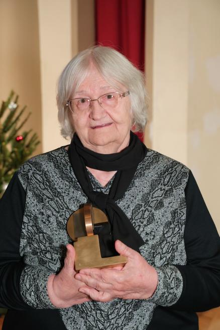 Divi cilvēki stāv uz skatuves zālē, sieviete rokās tur nelielu abstraktu metāla balvu, fonā redzams prezentācijas ekrāns.