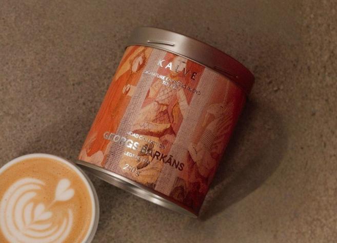 Uz pelēcīgas virsmas redzama kafijas bundža ar mākslinieciski noformētu etiķeti “KALVE x MDAD kolekcija, Georgs Barkāns” un blakus novietota kafijas tasīte ar latte art putu rakstu.