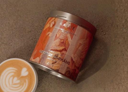 Uz pelēcīgas virsmas redzama kafijas bundža ar mākslinieciski noformētu etiķeti “KALVE x MDAD kolekcija, Georgs Barkāns” un blakus novietota kafijas tasīte ar latte art putu rakstu.