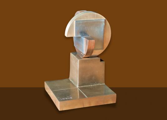 Rokās tiek turēts caurspīdīga stikla trauks, kas novietots uz dzintarkrāsas stikla plāksnes, izceļot materiālu caurspīdīgumu un faktūru.