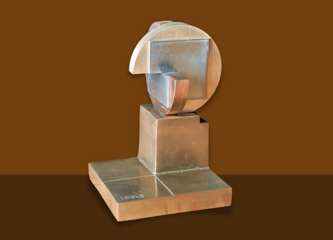 Rokās tiek turēts caurspīdīga stikla trauks, kas novietots uz dzintarkrāsas stikla plāksnes, izceļot materiālu caurspīdīgumu un faktūru.