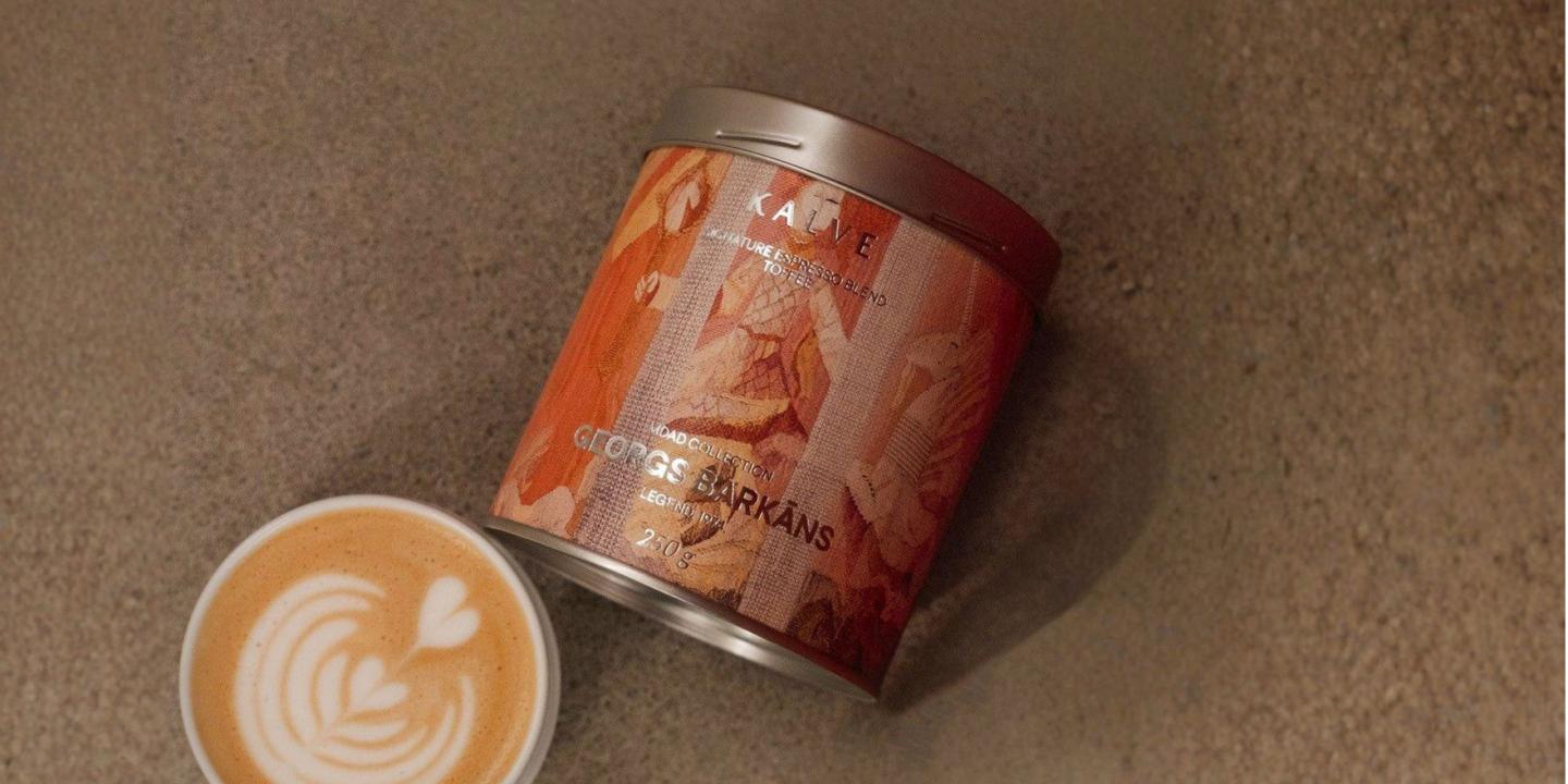 Uz pelēcīgas virsmas redzama kafijas bundža ar mākslinieciski noformētu etiķeti “KALVE x MDAD kolekcija, Georgs Barkāns” un blakus novietota kafijas tasīte ar latte art putu rakstu.