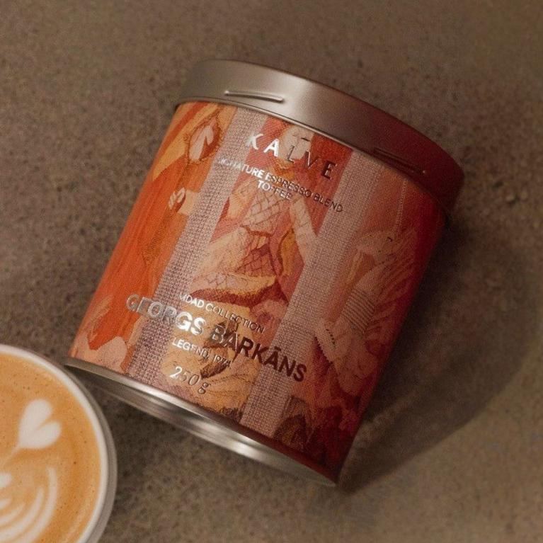 Uz pelēcīgas virsmas redzama kafijas bundža ar mākslinieciski noformētu etiķeti “KALVE x MDAD kolekcija, Georgs Barkāns” un blakus novietota kafijas tasīte ar latte art putu rakstu.