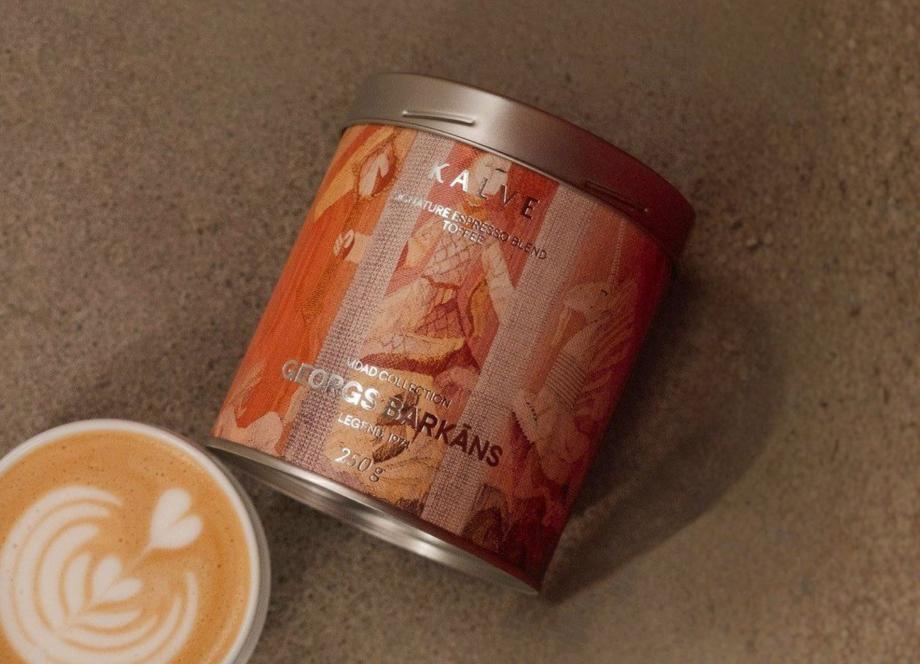 Uz pelēcīgas virsmas redzama kafijas bundža ar mākslinieciski noformētu etiķeti “KALVE x MDAD kolekcija, Georgs Barkāns” un blakus novietota kafijas tasīte ar latte art putu rakstu.