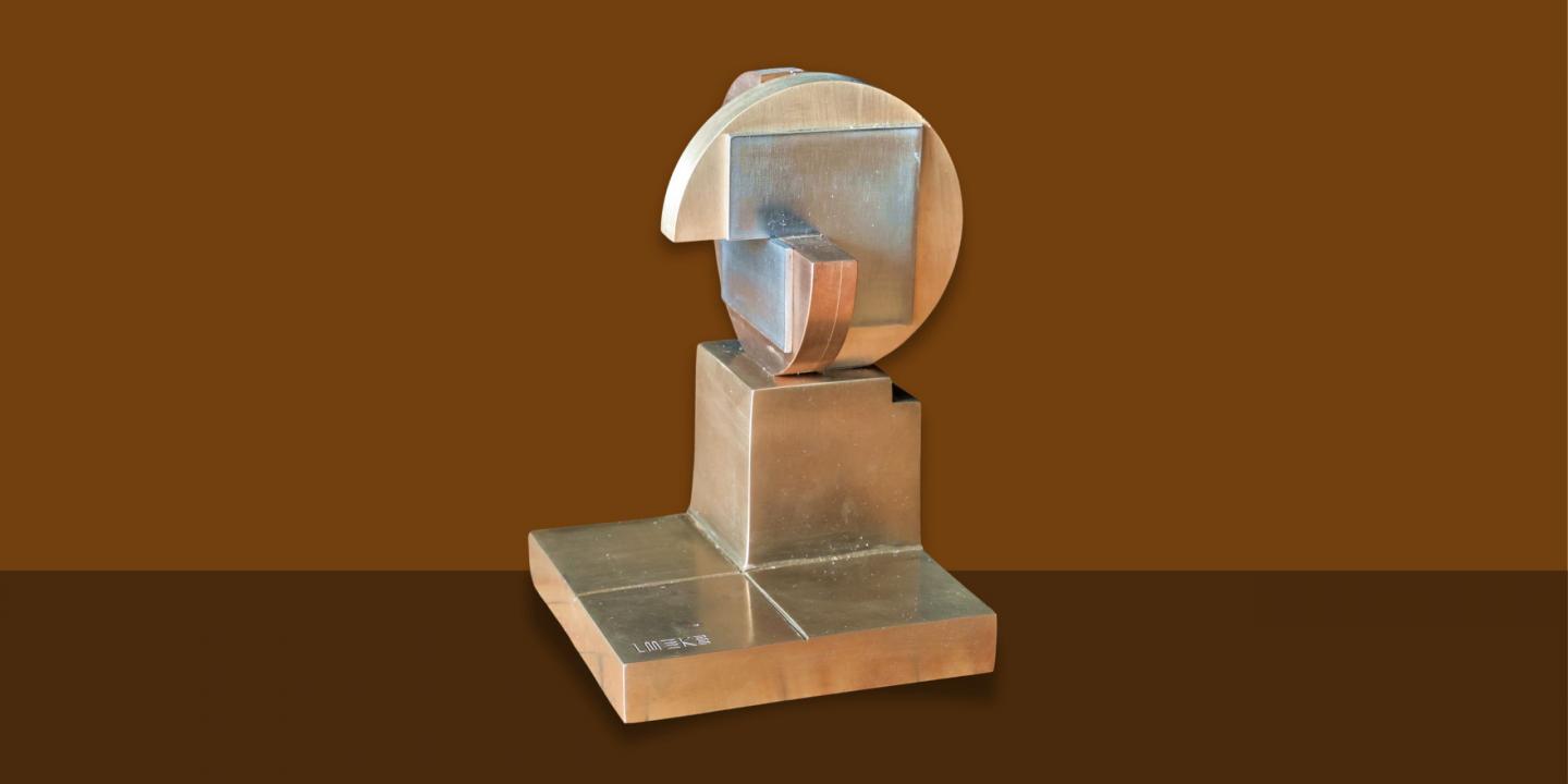 Metāla mākslas balva ar ģeometrisku, abstraktu formu uz kvadrātveida pamatnes, attēlota uz vienkrāsaina fona.
