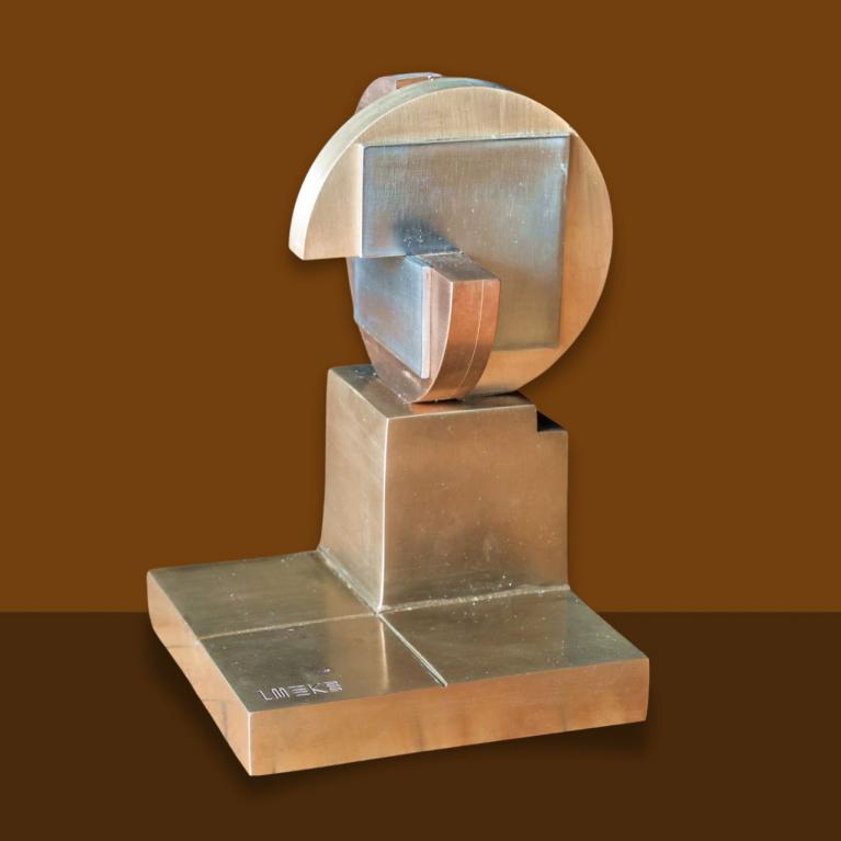 Metāla mākslas balva ar ģeometrisku, abstraktu formu uz kvadrātveida pamatnes, attēlota uz vienkrāsaina fona.