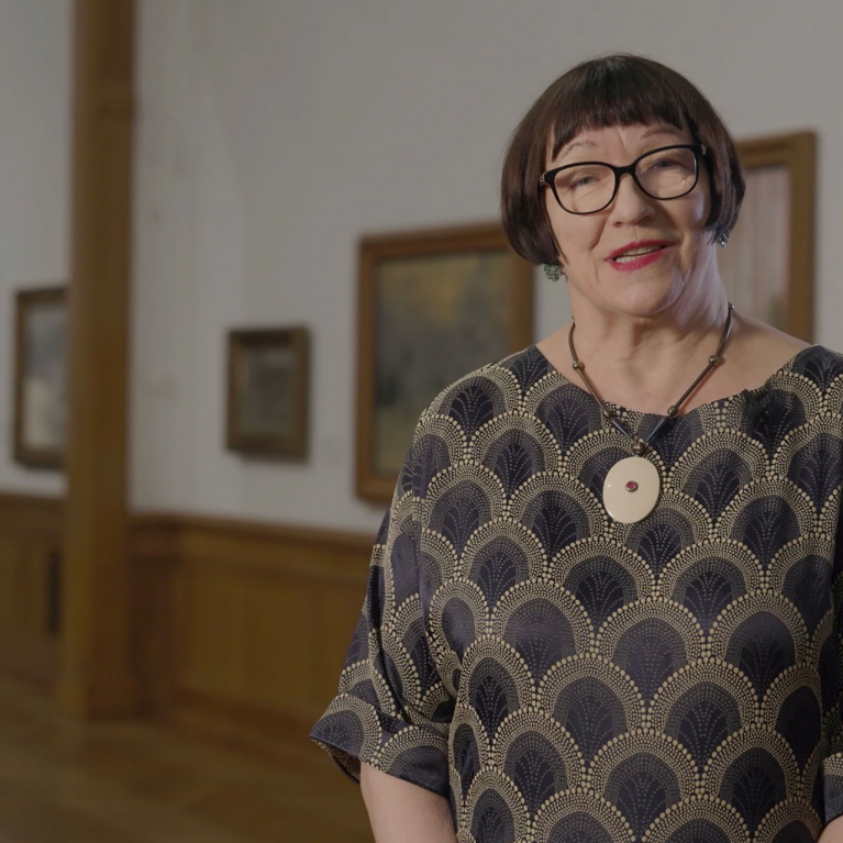 A woman wearing glasses and a patterned dress stands in the Latvian National Museum of Art exhibition hall, with framed paintings visible in the background.