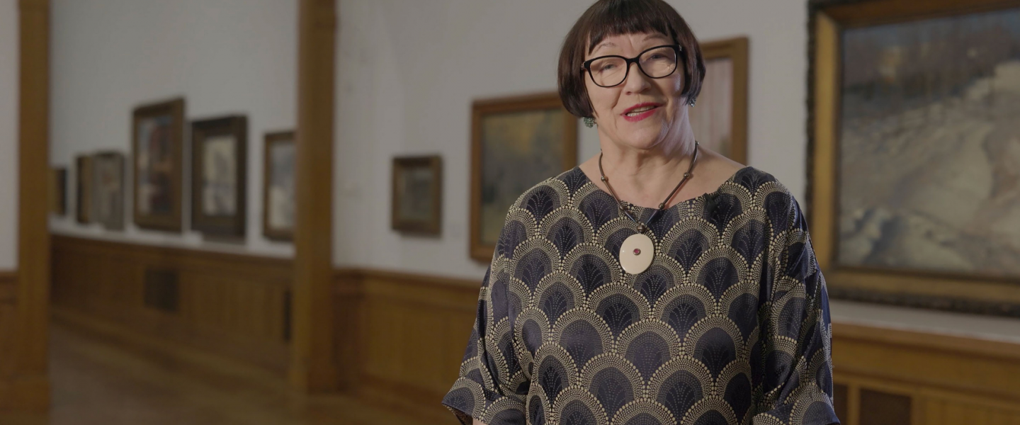 A woman wearing glasses and a patterned dress stands in the Latvian National Museum of Art exhibition hall, with framed paintings visible in the background.